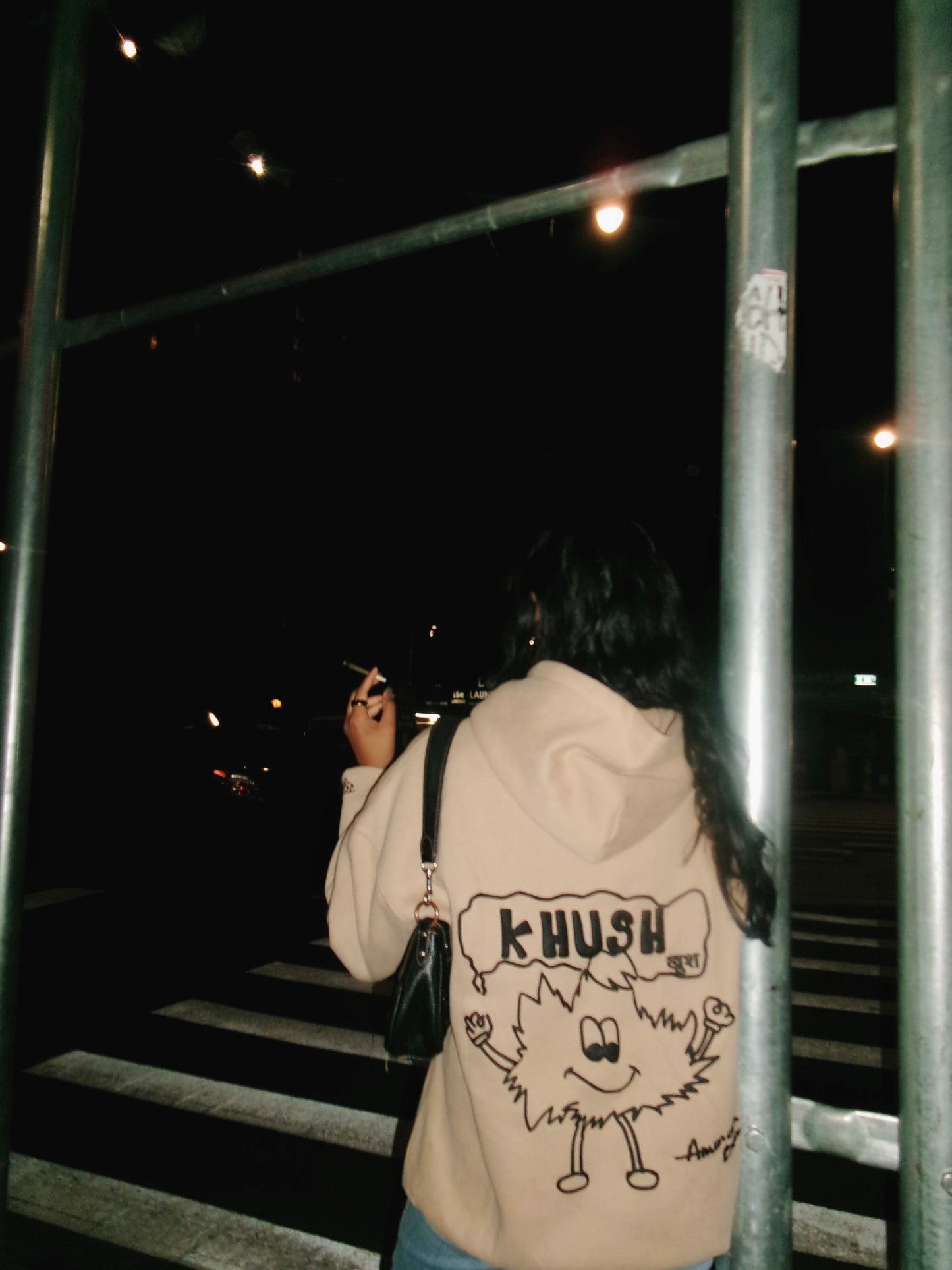A woman wearing a beige hoodie with a cartoon character and the text "KHUSH" on the back.