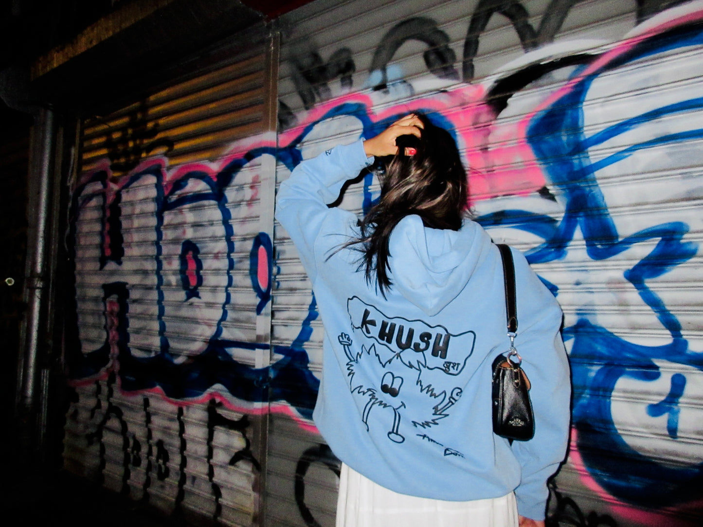 A woman in a light blue hoodie with a whimsical graphic on the back stands before colorful graffiti at night.